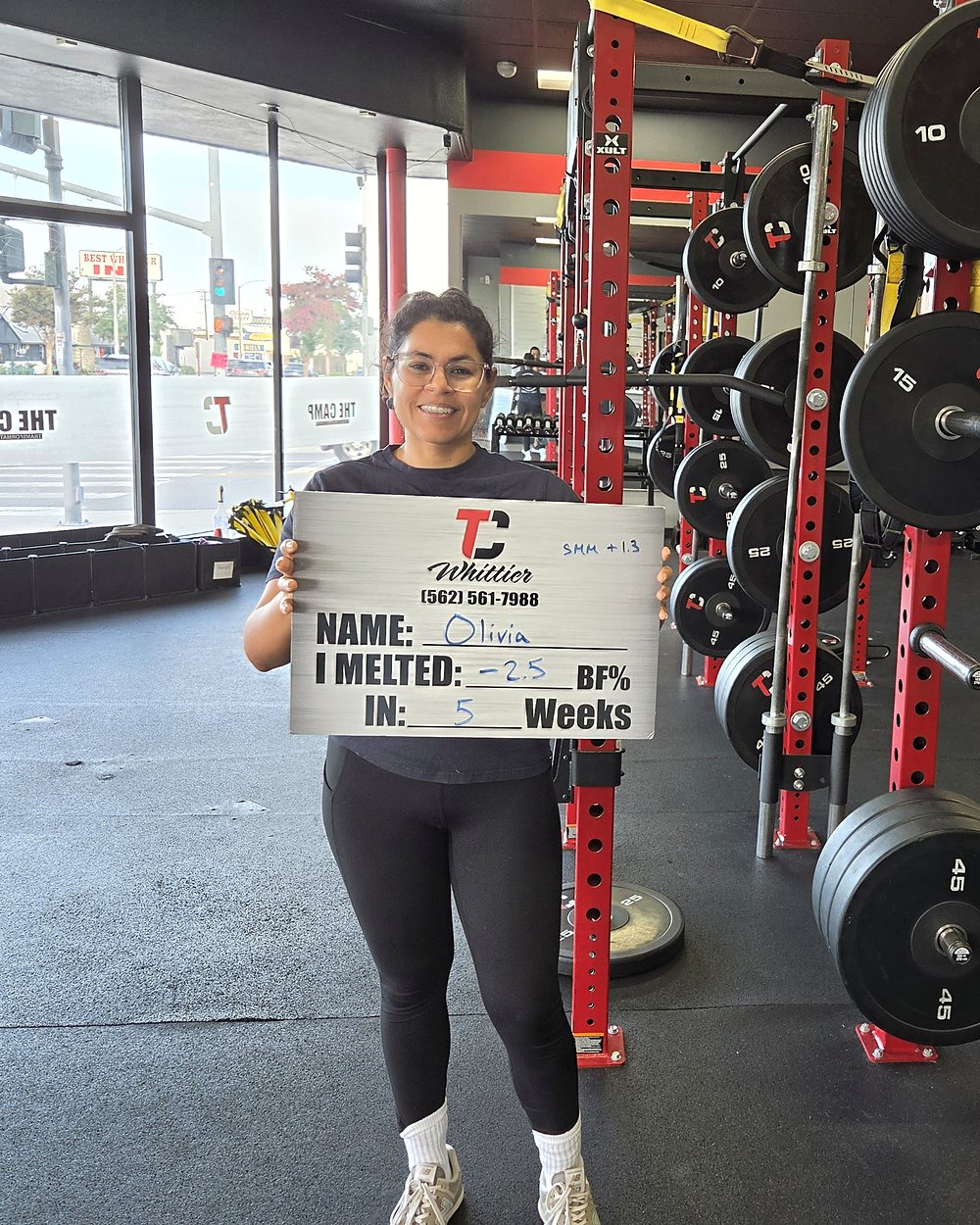 Olivia at a Camp Whittier weigh-in, holding a board that reads: melted 2.5 percent body fat in 5 weeks while adding muscle.